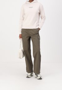 Sudadera beige claro con un logo, pantalones cargo verde oliva, bolso blanco y zapatillas estampadas. Conjunto casual con un corte holgado y detalles utilitarios.