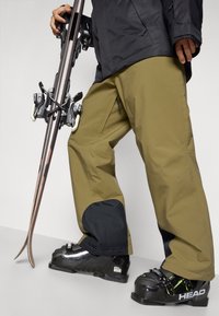 Pantalons de ski en vert olive avec des accents noirs, présentant une coupe décontractée, des genoux renforcés et des poches zippées sur les côtés, associés à des bottes de ski noires.