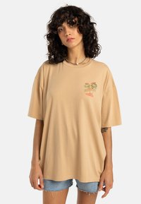 Beige oversized bomulls t-skjorte med korte ermer, med fargerik palmebroderi på venstre bryst. Blå denim shorts er synlige.