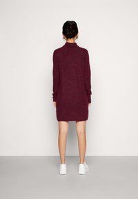 Bordeauxstickad klänning med hög krage, långa ärmar och avslappnad passform, bärs med vita sneakers. Slät textur med subtil värme.