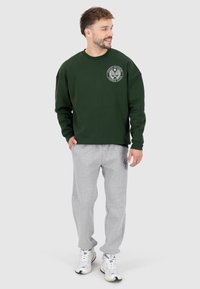 Felpa verde con dettagli a logo rotondo sul petto, abbinata a pantaloni tuta grigi con un logo circolare sulla coscia. Modello con scarpe da ginnastica bianche.