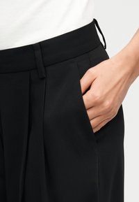 Χέρι που ακουμπά στην εμπρόσθια τσέπη μαύρου ψηλόμεσου παντελονιού με πιέτες, φορεμένο με λευκή ριμπ μπλούζα.