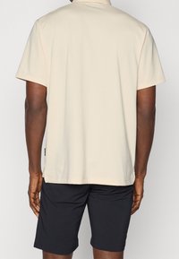 Polo beige à manches courtes avec un col, texture douce, fentes sur les côtés et shorts foncés. Le tissu semble léger et lisse.