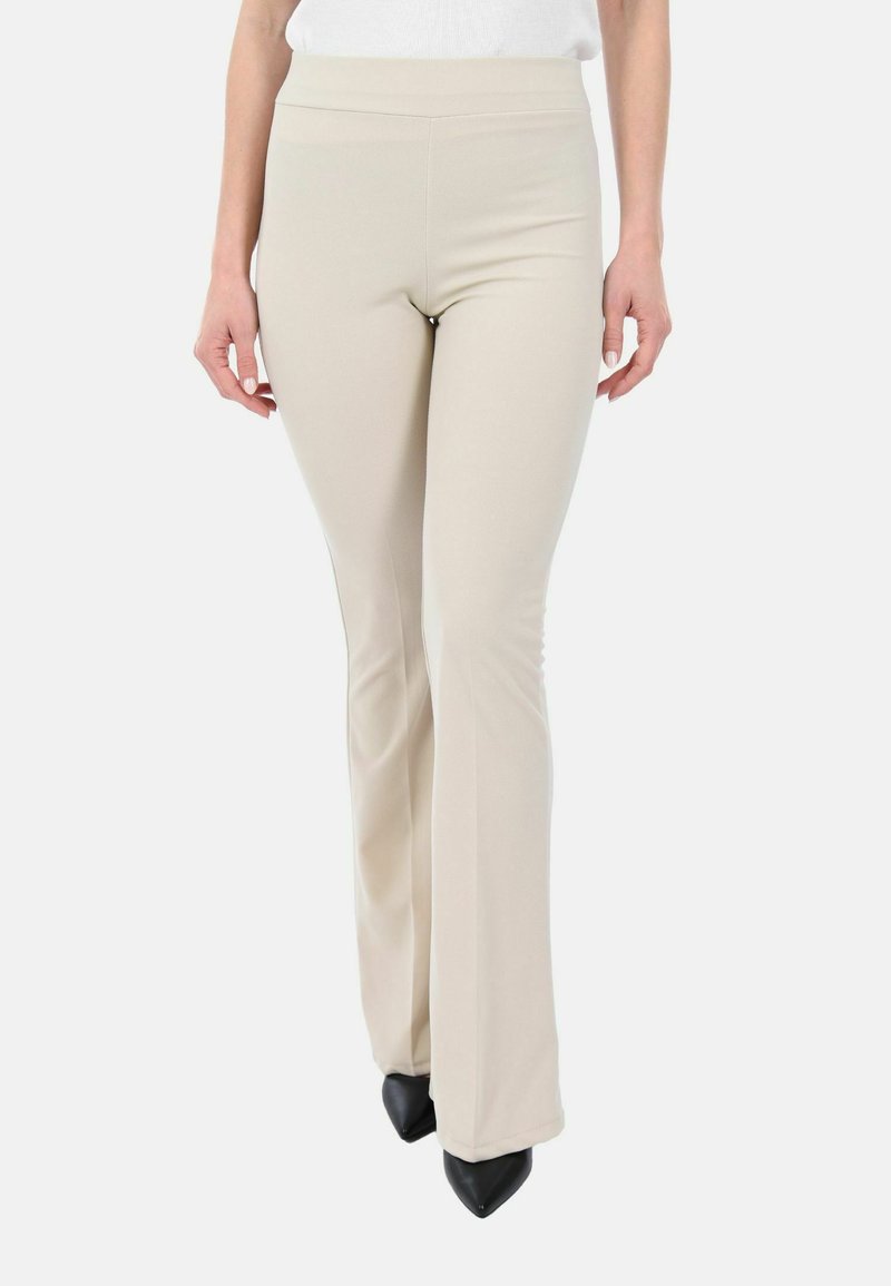 Donna che indossa pantaloni flare aderenti beige e scarpe nere con punta a punta, in piedi davanti a uno sfondo neutro.