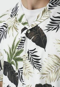 Camisa de manga corta en tela blanca con patrones de hojas tropicales en verde, negro y beige; incluye botones y un cuello desplegable.