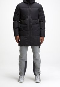 Doudoune noire avec plusieurs poches frontales, capuche ajustable et fermeture éclair à l'avant. Associée à un pantalon gris avec des accents latéraux noirs.