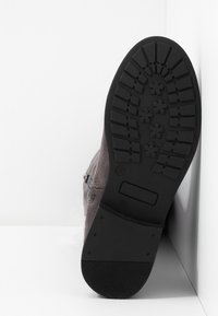 Šedá semišová bota s černou gumovou podrážkou s texturou, která má vroubkovaný vzor a boční zip pro snadné nazouvání.