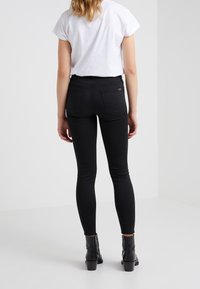 Nainen, jolla on yllään mustat slim fit -farkut, valkoinen lyhythihainen paita ja mustat nilkkurit, seisoo yksinkertaista valkoista taustaa vasten selkä kameraan päin.