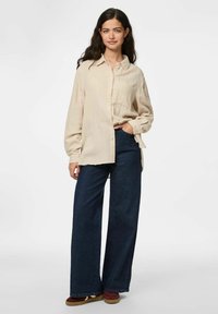 Camicia button-up beige chiaro con una finitura texture, abbinata a pantaloni in denim scuri a gamba larga e sneakers marroni.