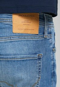 Blå denimjeans med en tan läderlapp med präglad text, standardfickor och synliga sömdetaljer för hållbarhet.