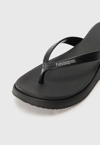 Svarte gummiflippflopper med strukturert såle, tynne stropper og logo på stroppen. Sålen har en lett kurve for komfort.