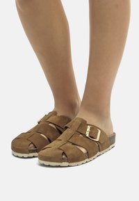 Sandales en suède marron avec un design tissé, dotées d'une boucle en or, ouvertes à l'avant et d'une semelle texturée pour une meilleure adhérence.
