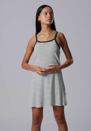 Pikkade juustega naine, kes kannab varrukateta valget kleiti mustade horisontaalsete triipudega, hoides käsi talje kõrgusel koos.