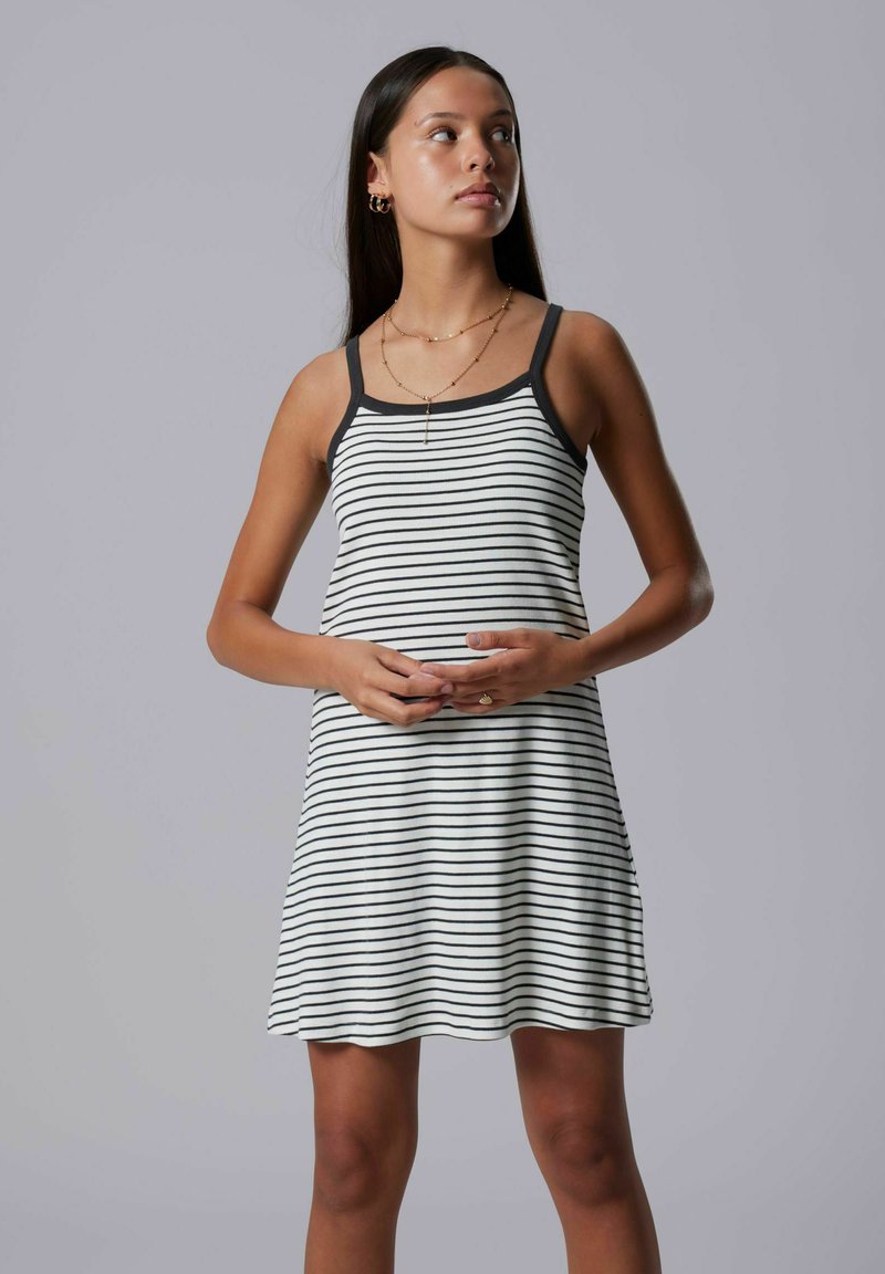 Femme aux longs cheveux portant une robe blanche sans manches à rayures horizontales noires, tenant ses mains ensemble au niveau de la taille.