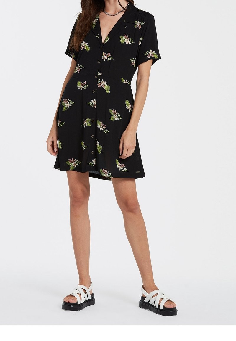 Black short-sleeve dress with floral print in green and pink. Features a button front and collar. Paired with white strappy sandals.