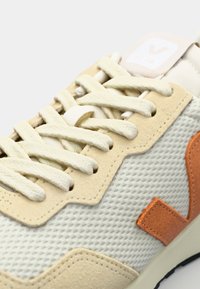 Lys beige sneakers laget med mesh og semsket skinn. Har teksturert overdel, flate snørebånd og en oransje sidologotype. Gummisåle.