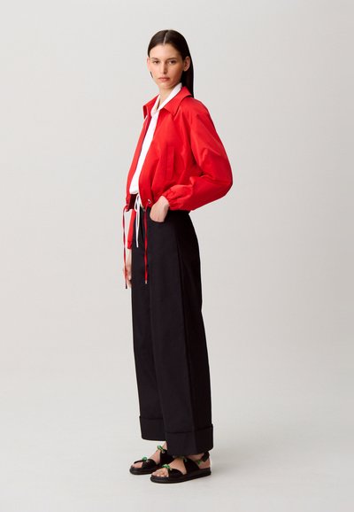 Giacca corta rossa, camicia bianca, pantaloni neri a gamba larga e sandali neri con accenti verdi. Tessuto liscio, vestibilità casual, design minimalista.