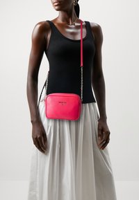 Różowa skórzana torebka na ramię o prostokątnym kształcie, z łańcuszkowym paskiem i subtelnym logo. Noszona z czarnym topem i białą spódnicą.