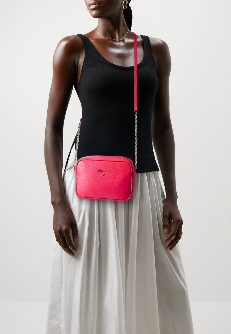 Różowa skórzana torebka na ramię o prostokątnym kształcie, z łańcuszkowym paskiem i subtelnym logo. Noszona z czarnym topem i białą spódnicą.