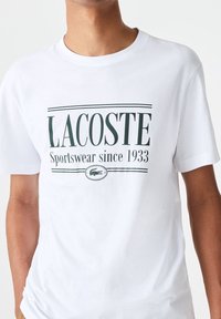Bílá bavlněná tílko se zeleným logem Lacoste a textem "Sportswear since 1933," s krátkým rukávem a klasickým kulatým výstřihem.