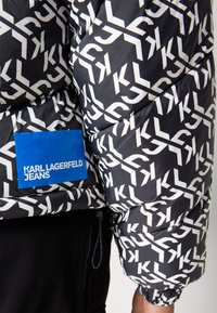 Kapitono striukė su juodos ir baltos spalvos logotipo raštu, mėlyna prekės ženklo etiketė ir elastinės rankovės. Pagaminta iš sintetinio pluošto su siuvinėjimo dizainu.