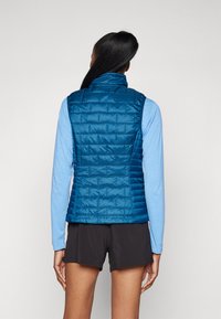 Gilet matelassé bleu avec un col haut, présentant une finition lisse et brillante, porté par-dessus une chemise à manches longues bleu clair et un short noir.