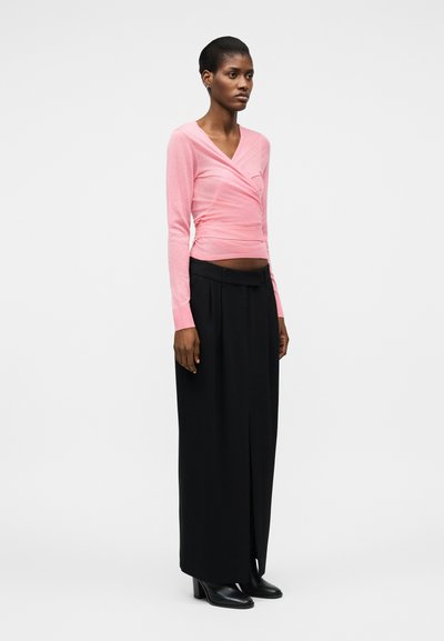 Femme debout portant un haut cache-cœur rose à manches longues, un pantalon large noir avec une fente à l'avant, et des bottes noires à talons sur un fond uni.