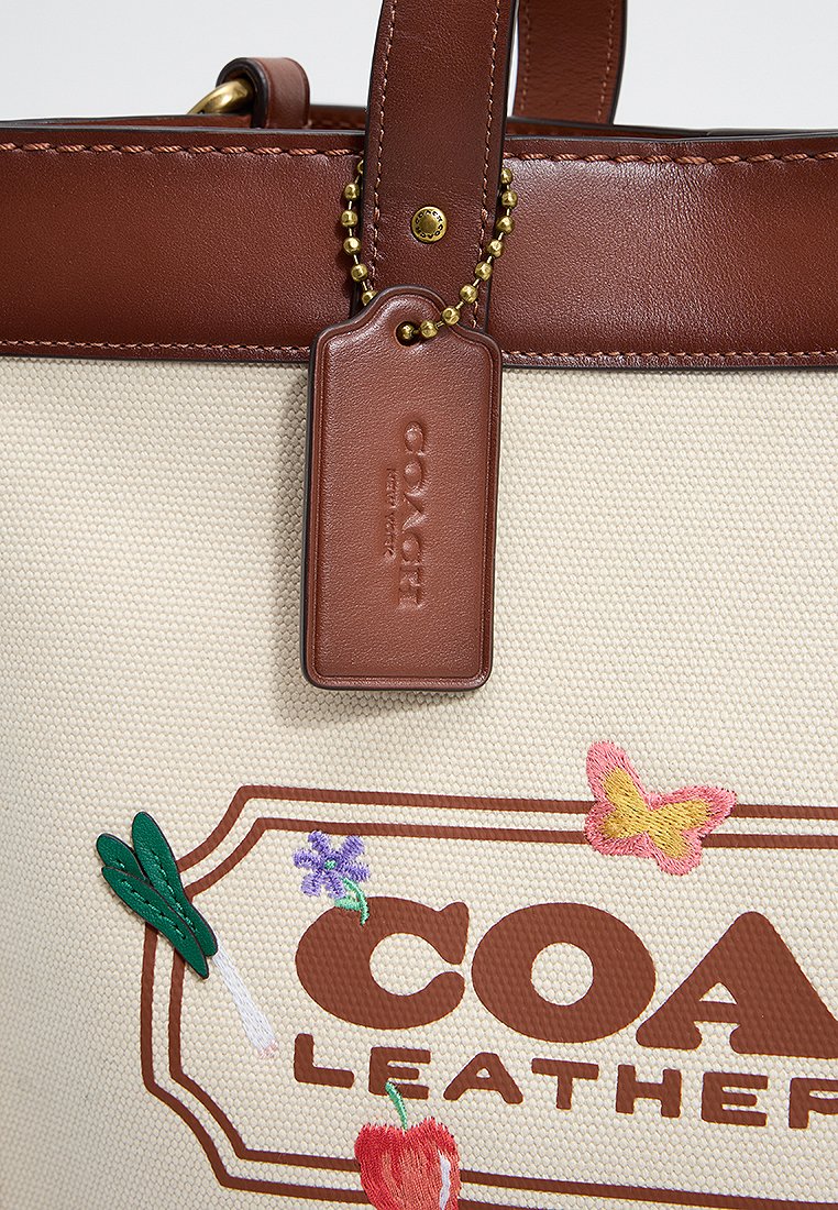 Bolso de lona color tawny con detalles de cuero marrón, que presenta bordados florales y una etiqueta de cuero con el logotipo de la marca.