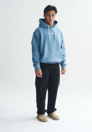 Light blue hoodie with a front pocket and white drawstrings, paired with black jeans and beige slip-on shoes.