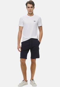 Maglietta bianca con un piccolo logo, pantaloni cargo blu navy con tasche laterali e sneakers bianche. L'outfit presenta un design casual e aderente.