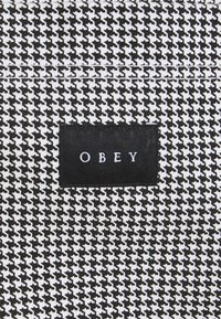 Tessuto a fantasia pied de poule bianco e nero con un'etichetta rettangolare nera su cui è scritto "OBEY" in bianco. Il tessuto ha un aspetto strutturato.