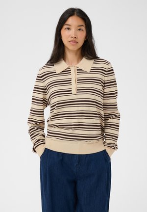 Kvinde med langt mørkt hår iført beige og brune stribede langærmet polo og mørkeblå bukser, stående med hænderne i lommerne.