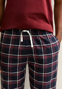 Man som bär en mörkröd skjorta och rutiga byxor i marinblått, röd och vitt med elastisk midja och vit dragsko, med händerna i fickorna.