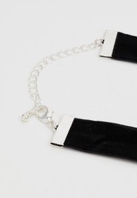 Choker de camurça preta com acabamentos em metal prateado, apresentando um fecho texturizado e uma extensão em corrente com fecho de clasp.