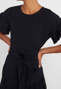 Schwarzes Kleid mit kurzen, gerafften Ärmeln, strukturiertem Stoff und einem gebundenen Taillenband. Verfügt über einen runden Ausschnitt und einen gestuften Rock.