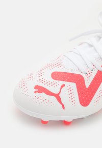 Scarpa da calcio bianca con accenti rosa e un motivo a pois. Presenta un logo prominente e una tomaia testurizzata, progettata per la presa e le prestazioni.