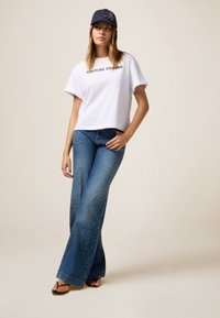 Witte katoenen t-shirt met de print "COUTURE CHARMÉ", gecombineerd met uitlopende donkerblauwe jeans met studs aan de zijkanten. Marineblauwe pet.