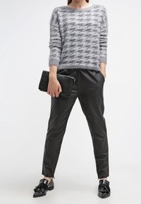 Donna che indossa un maglione grigio a quadri houndstooth, pantaloni in pelle neri, mocassini neri con frange, tiene una pochette nera con una mano in tasca.