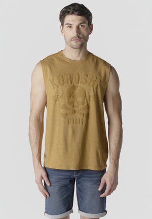 Hombre con cabello corto y oscuro, vistiendo una camiseta sin mangas mostaza con diseño de calavera y shorts de mezclilla, de pie frente a un fondo claro y liso.