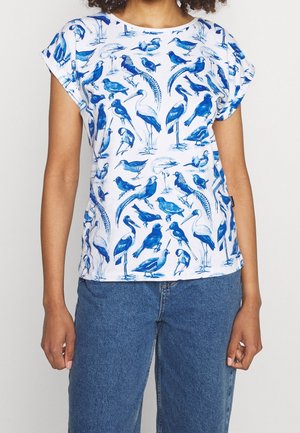 Femme portant une chemise blanche à manches courtes avec des illustrations d'oiseaux bleus et un jean bleu, debout avec les bras détendus le long du corps.