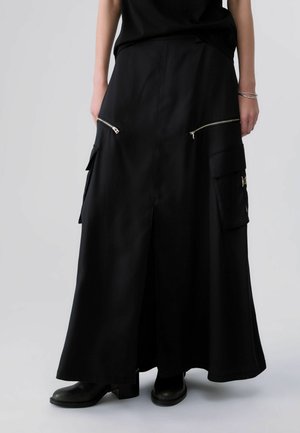 Pantalon noir à jambes larges avec grandes poches zippées et à rabats, porté avec des bottes noires et un haut noir sans manches.