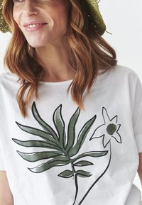 White short-sleeve shirt featuring green botanical illustrations of leaves and a flower, with a round neckline and relaxed fit.