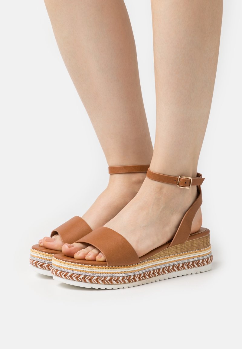 Pieds portant des sandales compensées marron avec des brides à la cheville et des semelles tissées aux motifs blancs, marrons et beiges sur fond blanc.