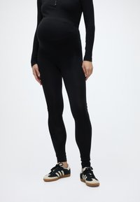 Έγκυος που φοράει μαύρο παντελόνι εγκυμοσύνης, μαύρο μακρυμάνικο μπλουζάκι και μαύρα-άσπρα αθλητικά παπούτσια, στέκεται μπροστά σε απλό φόντο.