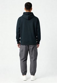 Uomo che indossa un hoodie nero, pantaloni cargo grigi e sneakers bianche, in piedi di spalle contro uno sfondo chiaro e semplice.