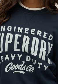 Sudadera azul marino de tela suave con un gran texto impreso en blanco que dice "ENGINEERED SUPERDRY HEAVY DUTY Goods Co." y un cuello en blanco.
