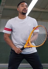 Grå t-skjorte med korte ermer og røde striper, kombinert med svarte bukser. Holder en gul tennisracket med svart grep på en bane.