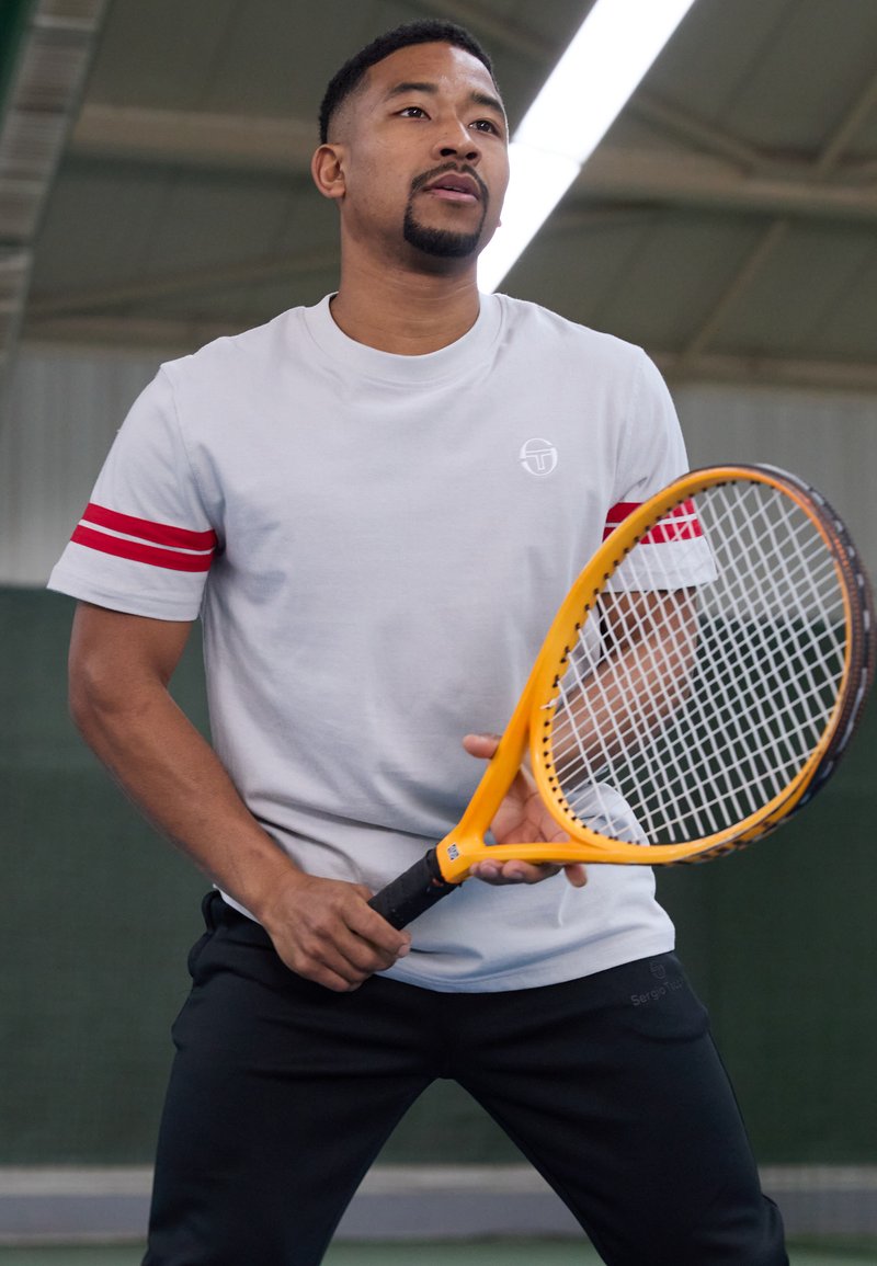 Grå t-skjorte med korte ermer og røde striper, kombinert med svarte bukser. Holder en gul tennisracket med svart grep på en bane.