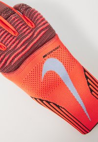 Röd och svart atletisk handske med texturerat grepp, har en stickad ovandel och Nike-logotyp i vitt. Förstärkta fingertoppar och elastisk mudden.