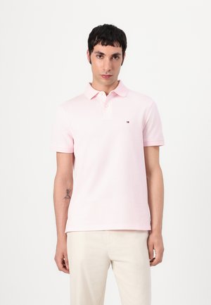 Polo en coton rose clair avec manches courtes, un col et un petit logo sur la poitrine gauche. Texture lisse, coupe décontractée.